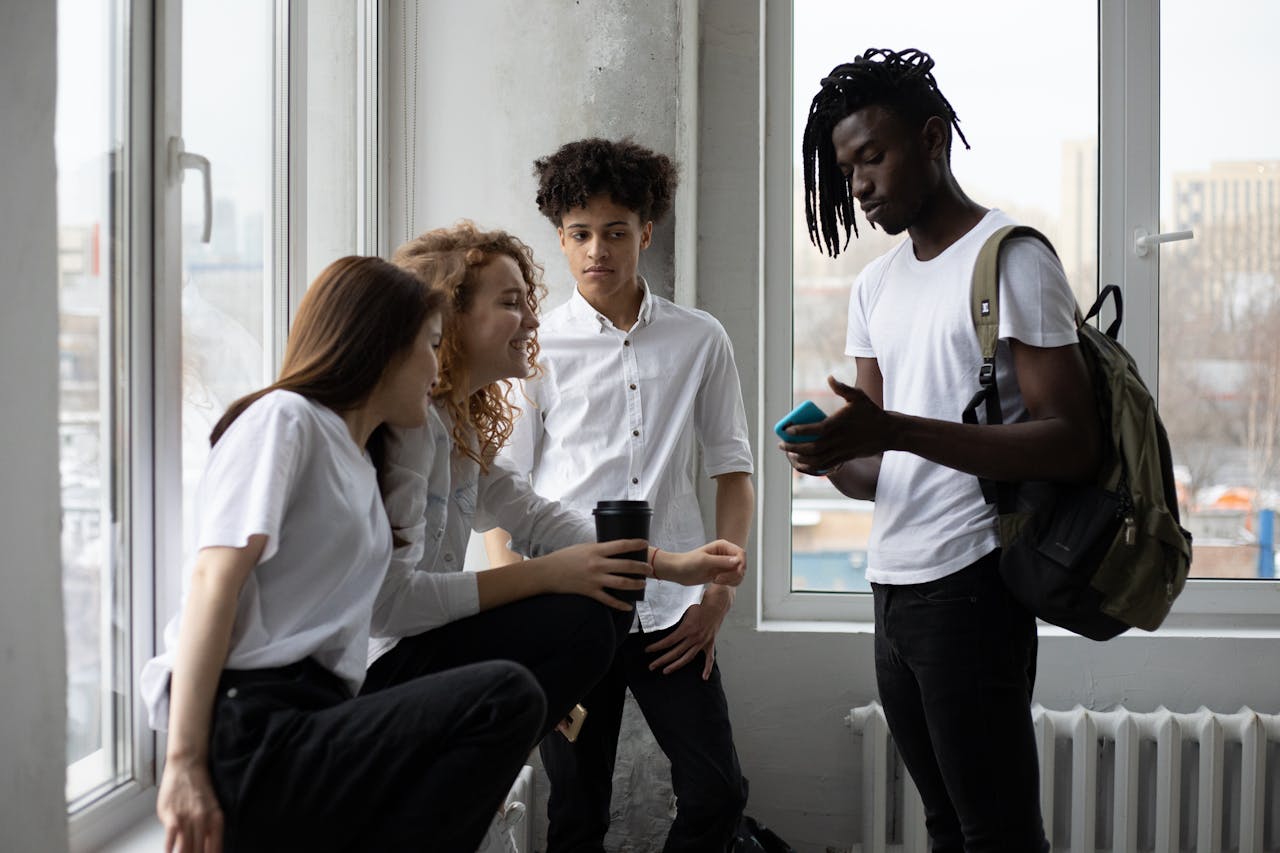 hero-img-01 Group of multiracial classmates with backpack and takeaway coffee having conversation at windowsill near window in light classroom before lesson