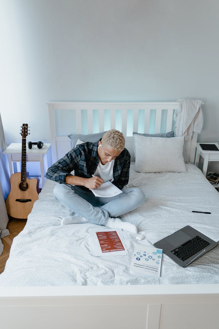 about-01 A young man focuses on his studies, sitting on a bed with books and a laptop in a cozy bedroom.
